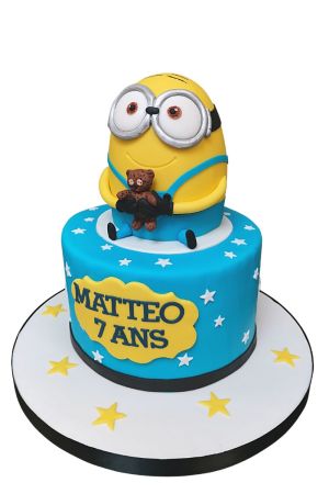 Exclusieve blauwe verjaardagstaart met witte sterren, bekroond met een handgemaakt 3D Minion figuurtje dat een teddybeer vasthoudt, van The French Cake Company.