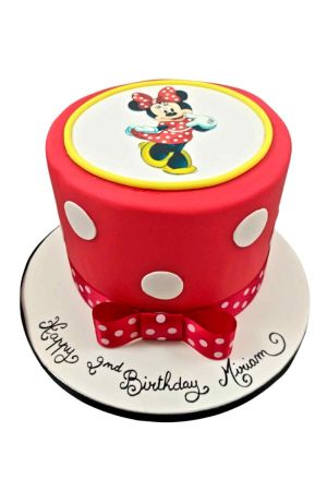 Un gâteau d'anniversaire rond en pâte à sucre rouge à pois blancs avec un ruban et un nœud rouge à pois blancs à la base, surmonté d'une impression alimentaire ronde représentant Minnie Mouse entourée d'un liseré jaune.