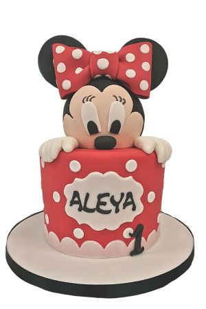 A round red fondant birthday cake with white polka dots, decorated on the front with a white scalloped plaque and a black number one, topped with a large 3D sugar model of Minnie Mouse peeking over the top edge with her hands, wearing a large red and whit