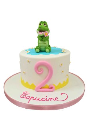 Un gâteau d'anniversaire rond blanc en pâte à sucre décoré de petites fleurs, surmonté d'une figurine de crocodile vert tenant une fleur rose devant un grand chiffre deux rose.