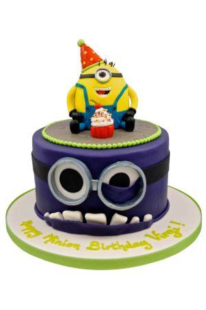 Exclusieve paarse Evil Minion monster verjaardagstaart met grote duikbril en tanden, bekroond met een gele Minion met feesthoedje bij een cupcake, van The French Cake Company.