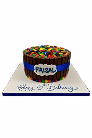 Un gâteau d'anniversaire rond entouré de barres KitKat en chocolat, noué d'un ruban bleu et recouvert d'une montagne de bonbons M&M's colorés.