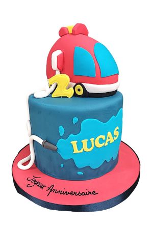 Un gâteau d'anniversaire rond en pâte à sucre bleu foncé orné d'une tache d'eau bleu clair, surmonté d'un grand camion de pompier rouge modelé en 3D, d'un chiffre deux jaune et d'un tuyau d'incendie blanc.