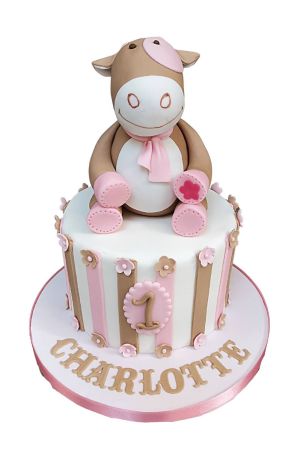 Witte 1e verjaardagstaart met roze en beige verticale strepen, kleine suikerbloemetjes, een ovaal schildje met cijfer 1 en een grote 3D handgemaakte Noukies Lola de koe figuur gemaak door The French Cake Company