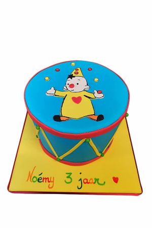 Gâteau d'anniversaire bleu en forme de tambour de cirque avec cordelettes jaunes, bordure rouge et illustration de Bumba le clown sur un socle jaune vif réalisé par The French Cake Company