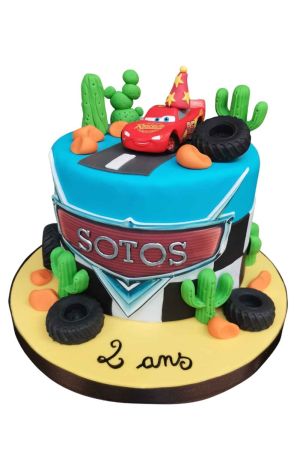 Gâteau d'anniversaire bleu turquoise sur le thème de Flash McQueen avec figurine 3D sur route en bitume, cactus en pâte à sucre, rochers et pneus de course réalisé par The French Cake Company