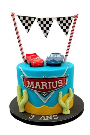 Gâteau d'anniversaire bleu ciel Disney Cars décoré de figurines de Flash McQueen et Finn McMissile, cactus en sucre, dunes du désert et guirlande à fanions damier par The French Cake Company
