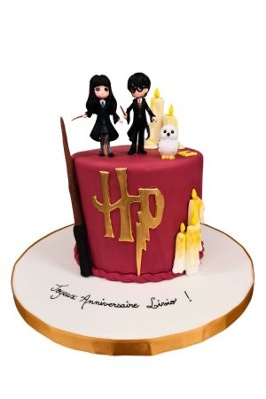 Un gâteau d'anniversaire rond en pâte à sucre bordeaux décoré du logo doré HP et d'une grande baguette magique sur le côté, surmonté de figurines d'Harry Potter, d'Hermione Granger, de la chouette blanche Hedwige et de bougies fondues en sucre.