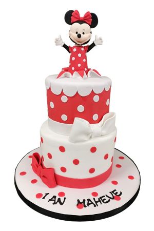 Une pièce montée d'anniversaire à deux étages en pâte à sucre blanche à pois rouges et rouge à pois blancs, ornée de nœuds en sucre et surmontée d'une figurine 3D de Minnie Mouse.