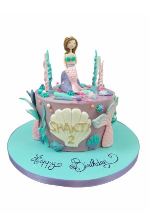 A purple and turquoise birthday cake featuring a handcrafted 3D mermaid sitting on a rock, decorated with edible seashells, coral, and a personalized gold shell.