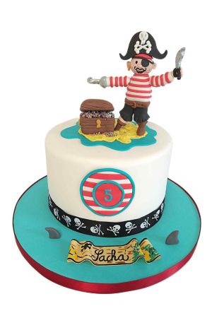 A round white fondant birthday cake decorated with a skull-patterned border, topped with a 3D sugar figurine of a pirate holding a sword next to an open treasure chest on a sandy island.