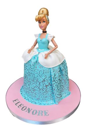 Assepoester pop verjaardagstaart met een blauwe fondant baljurk, zilveren krullen en een witte schoot op een roze taartplateau gemaakt door The French Cake Company