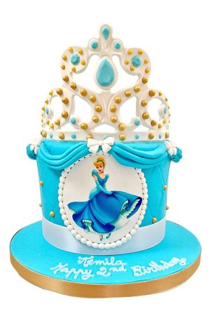 Hemelsblauwe gewatteerde prinsessen verjaardagstaart met Assepoester cameo ovaal, witte gedrapeerde linten en een grote witte met gouden diadeem topper gemaakt door The French Cake Company