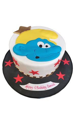 Un gâteau d'anniversaire rond blanc orné du visage d'un Schtroumpf bleu au chapeau jaune en pâte à sucre, entouré d'étoiles rouges sur un socle noir.