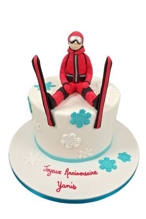 Un gâteau d'anniversaire blanc décoré de flocons turquoise et blancs, surmonté d'une figurine 3D d'un skieur en combinaison rouge avec ses skis.