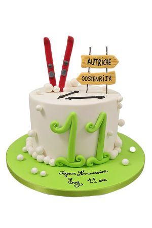 A white birthday cake decorated with lime green numbers, red edible skis, white fondant snowballs, and wooden-style trail signs saying Autriche and Oostenrijk.