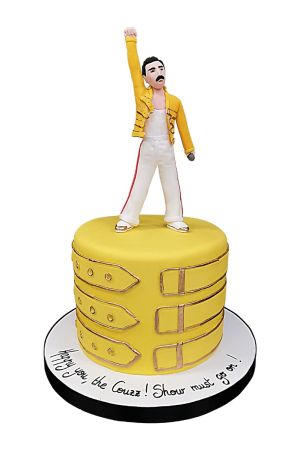 Un gâteau d'anniversaire rond en pâte à sucre jaune vif arborant des lanières de boucles dorées, surmonté d'une figurine 3D en sucre de Freddie Mercury avec sa veste jaune emblématique et le poing levé vers le ciel.
