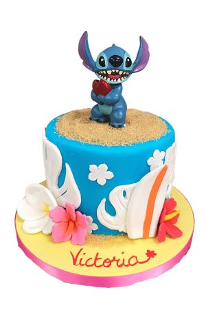 A blue fondant birthday cake with a 3D Stitch figure on edible sand, decorated with white tropical leaves, a surfboard, and pink hibiscus flowers.