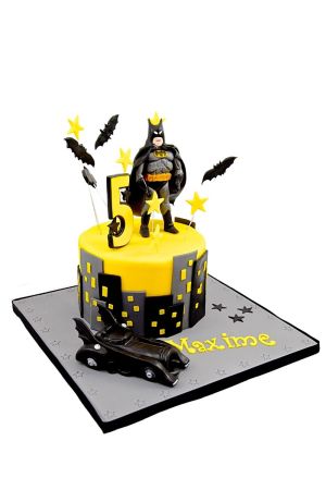 Yellow round superhero birthday cake decorated with black Gotham City skyscraper cutouts, flying bat toppers, a Batman figurine, and a black Batmobile car on a gray star-embossed board by The French Cake Company