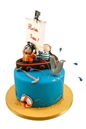 A round blue fondant birthday cake decorated with a silver anchor and lifebuoy, topped with a detailed 3D brown sailboat carrying two sugar pirate figurines next to a large grey whale tail splashing out of the water.