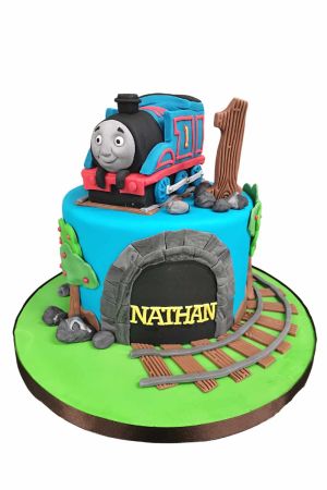 Gâteau du 1er anniversaire bleu turquoise sur plateau vert représentant un tunnel en pierre, des rails en bois courbés avec rochers en sucre, un chiffre 1 effet bois et la locomotive 3D Thomas le Petit Train au sommet réalisé par The French Cake Company