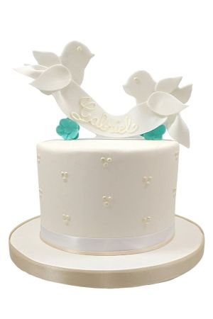 Gâteau de baptême blanc pour garçon à un étage, décoré de points de nacre, de fleurs turquoise et de deux colombes tenant une bannière.