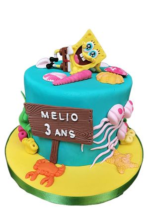 Bright turquoise SpongeBob SquarePants birthday cake featuring a relaxing SpongeBob topper, fondant jellyfish, orange crab, starfish, shells, and wooden sign on a yellow sand cake board by The French Cake Company