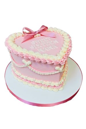 Un gâteau d'anniversaire haut en forme de cœur rose, décoré dans un style Lambeth vintage avec des bordures de crème blanches et roses festonnées, des perles comestibles et un nœud en satin rose.