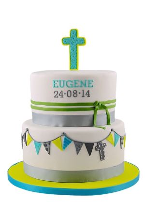 A two-tier white tiered communion cake decorated with silver ribbons, a lime green cross topper, and colorful hanging bunting.