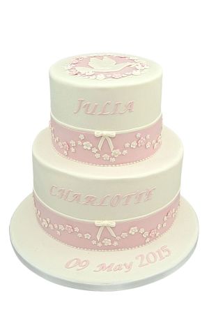 A two-tier white and pink communion cake for a girl, decorated with sugar flowers, ribbons, and a white dove of peace on the top tier.