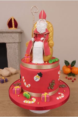 A two-tiered festive Saint Nicholas cake on a red cake stand, featuring a figurine of a bishop (Saint Nicholas) seated on a golden throne, holding a crozier. The cake is decorated with small gifts, candy canes, snowmen, and gingerbread cookies. The backgr