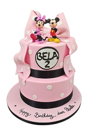 A two-tier pink and white birthday cake for a 1st birthday, decorated with white polka dots, a quilted pattern, a large pink sugar bow, and black Minnie Mouse ears on top.