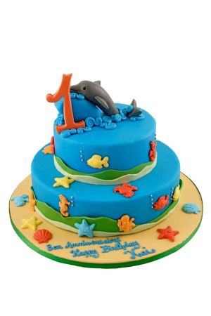 Un grand gâteau d'anniversaire bleu à deux étages sur le thème sous-marin, décoré de poissons et d'étoiles de mer colorés, surmonté d'un dauphin gris en sucre et d'un chiffre un orange.