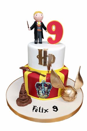 Une pièce montée d'anniversaire à deux étages en pâte à sucre blanche et rayée rouge et jaune Gryffondor, surmontée d'un petit personnage de sorcier en sucre et d'un chiffre neuf, décorée avec le blason de la maison, un Choixpeau, un balai et un Vif d'Or.