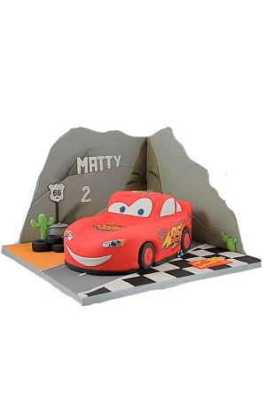 Elaborate 3D sculpted red Lightning McQueen car birthday cake set against a backdrop of sugar mountains, Route 66 sign, cacti, stack of tires, and a checkered race track board by The French Cake Company