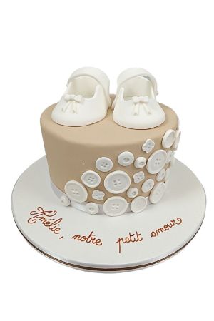 Gâteau de baptême beige à un étage, décoré de boutons blancs en pâte à sucre et surmonté de chaussons blancs artisanaux.