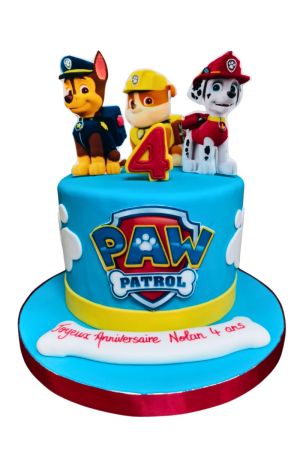 Een ronde lichtblauwe fondant verjaardagstaart met het Paw Patrol logo en witte hondenpootjes, bekroond met figuren van Chase, Rubble en Marshall en een rood cijfer 4 op een taartkarton met naam.