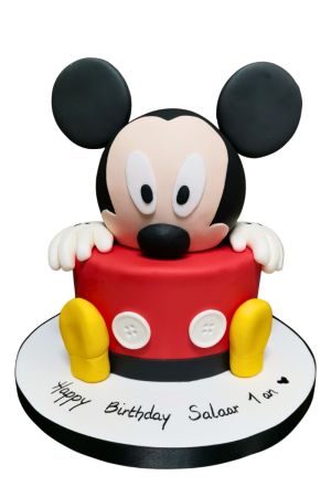 Gâteau d'anniversaire sculpté en 3D représentant Mickey Mouse dépassant d'un gâteau rouge orné de ses boutons blancs et grands souliers jaunes, créé par The French Cake Company.