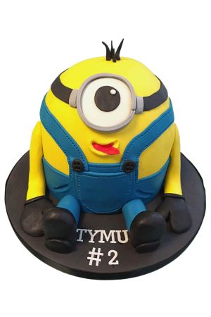 Gâteau d'anniversaire sculpté en 3D représentant un Minion jaune assis vêtu d'une salopette en jean bleue avec son œil unique, créé par The French Cake Company.
