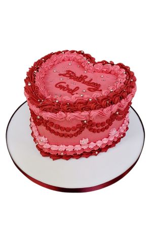 Un gâteau d'anniversaire en forme de cœur rose, décoré dans un style Lambeth vintage avec des bordures de crème festonnées roses et rouge foncé.