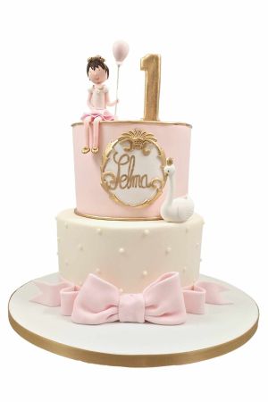 Luxe twee-laags roze en crème 1e verjaardagstaart met een geboetseerd meisje met ballon, een koninklijke zwaan en gouden getal 1 gemaak door The French Cake Company