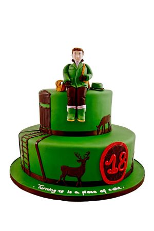 Two-tier green hunting birthday cake featuring a sculpted hunter figurine sitting on top, wooden watchtower ladder, and stag silhouettes