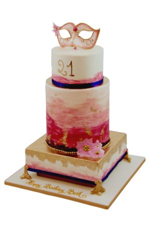Une pièce montée de luxe à trois étages décorée d'un effet aquarelle rose, de feuilles d'or et d'un masque vénitien rose fait main au sommet.
