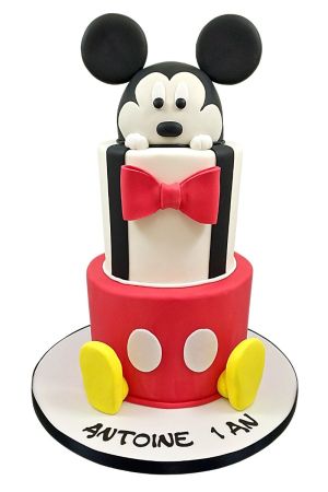 Gâteau d'anniversaire personnalisé à deux étages Mickey Mouse arborant un pantalon rouge à boutons, bretelles noires, nœud papillon et tête sculptée, créé par The French Cake Company.