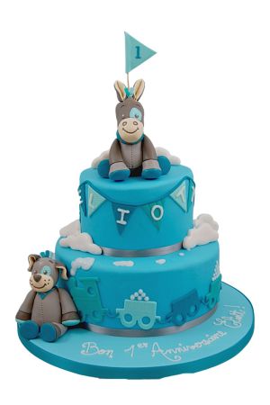 Blauwe twee-laags 1e verjaardagstaart met vlaggetjes, een stoomtreintje rond de basis, wolkjes en 3D geboetseerde figuren van Noukies Cyril de ezel en Gaston de hond gemaakt door The French Cake Company