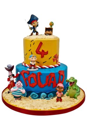 A two-tiered birthday cake covered in yellow and blue fondant, designed like a treasure island with edible sand and waves, featuring toys of Jake and the Never Land Pirates characters around it.