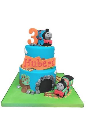 Pièce montée d'anniversaire à deux étages bleue sur plateau vert, avec rails de chemin de fer en bois, entrée de tunnel en pierre et figurines 3D de Thomas le Petit Train et Percy réalisée par The French Cake Company