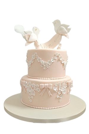 Gâteau de baptême à deux étages rose pâle décoré de fleurs blanches en sucre, de nœuds et de deux colombes tenant une bannière.