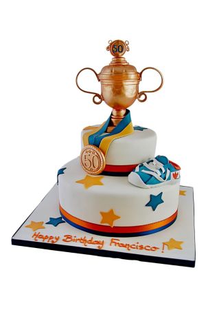 A two-tiered white fondant birthday cake decorated with blue and orange sugar stars, topped with a large golden trophy cup, a gold medal, and a small blue and white sports shoe.