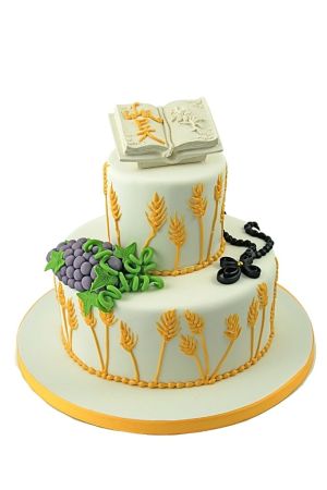 A two-tier white communion cake decorated with golden wheat stalks, purple grapes, a black rosary, and an open sugar Bible on top.